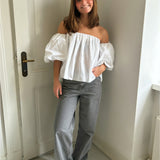 En person står afslappet i et rum med lyse vægge og en hvid dør. Personen bærer en hvid off-shoulder-bluse og grå bukser, og de smiler.