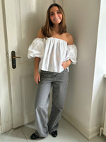 En person står afslappet i et rum med lyse vægge og en hvid dør. Personen bærer en hvid off-shoulder-bluse og grå bukser, og de smiler.