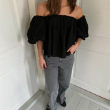 En kvinde står ved en lys væg iført en sort off-shoulder bluse med pufærmer, grå jeans og sorte sko. Hun har hånden i lommen og rører ikke ved væggen.