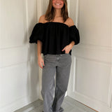 En person står afslappet iført en sort off-shoulder bluse og grå denim bukser i et rum med lyse vægge og gulve.