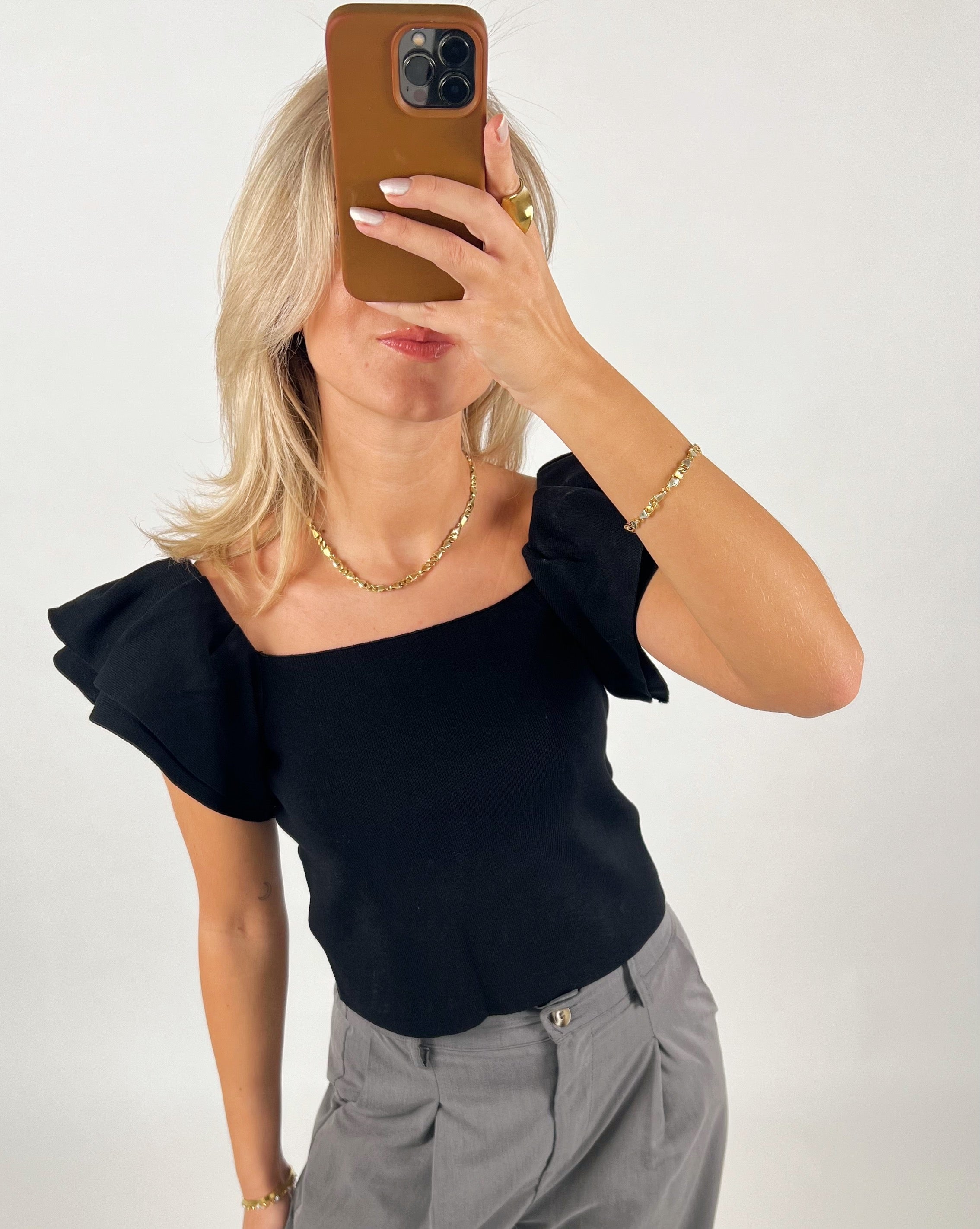 En kvinde tager et spejlbillede af sig selv med en telefon. Hun har en sort bluse med pufærmer og grå bukser på en hvid baggrund.