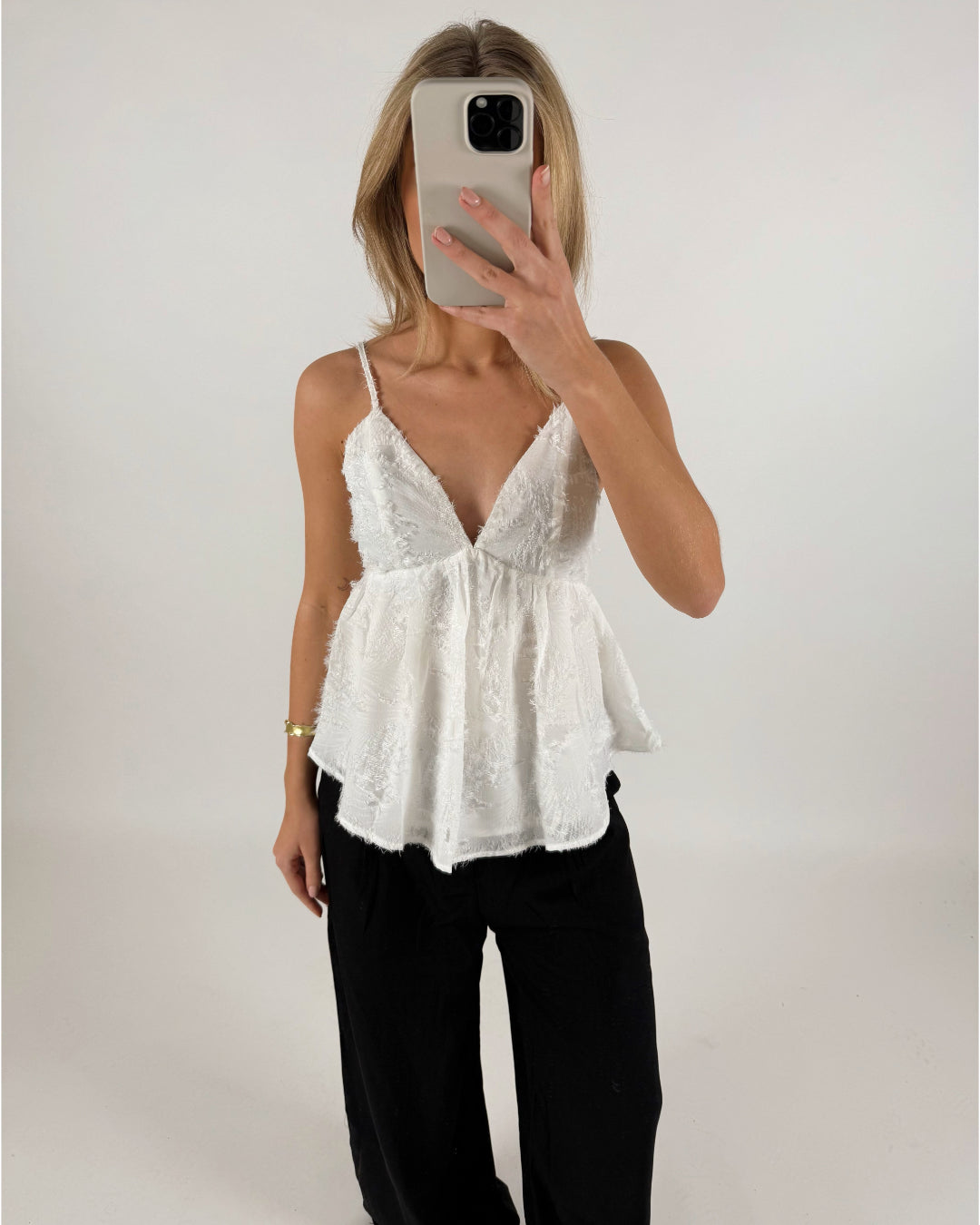 En person iført en hvid bluse med tynde stropper holder en mobiltelefon foran ansigtet. Personen står i et enkelt, lyst rum.