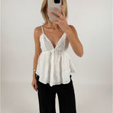 En person iført en hvid bluse med tynde stropper holder en mobiltelefon foran ansigtet. Personen står i et enkelt, lyst rum.