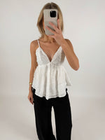 En person iført en hvid bluse med tynde stropper holder en mobiltelefon foran ansigtet. Personen står i et enkelt, lyst rum.