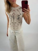 En person i en lysebrun, mønstret striktop og hvide bukser tager et spejlbillede med en smartphone, i et enkelt, lyst rum.