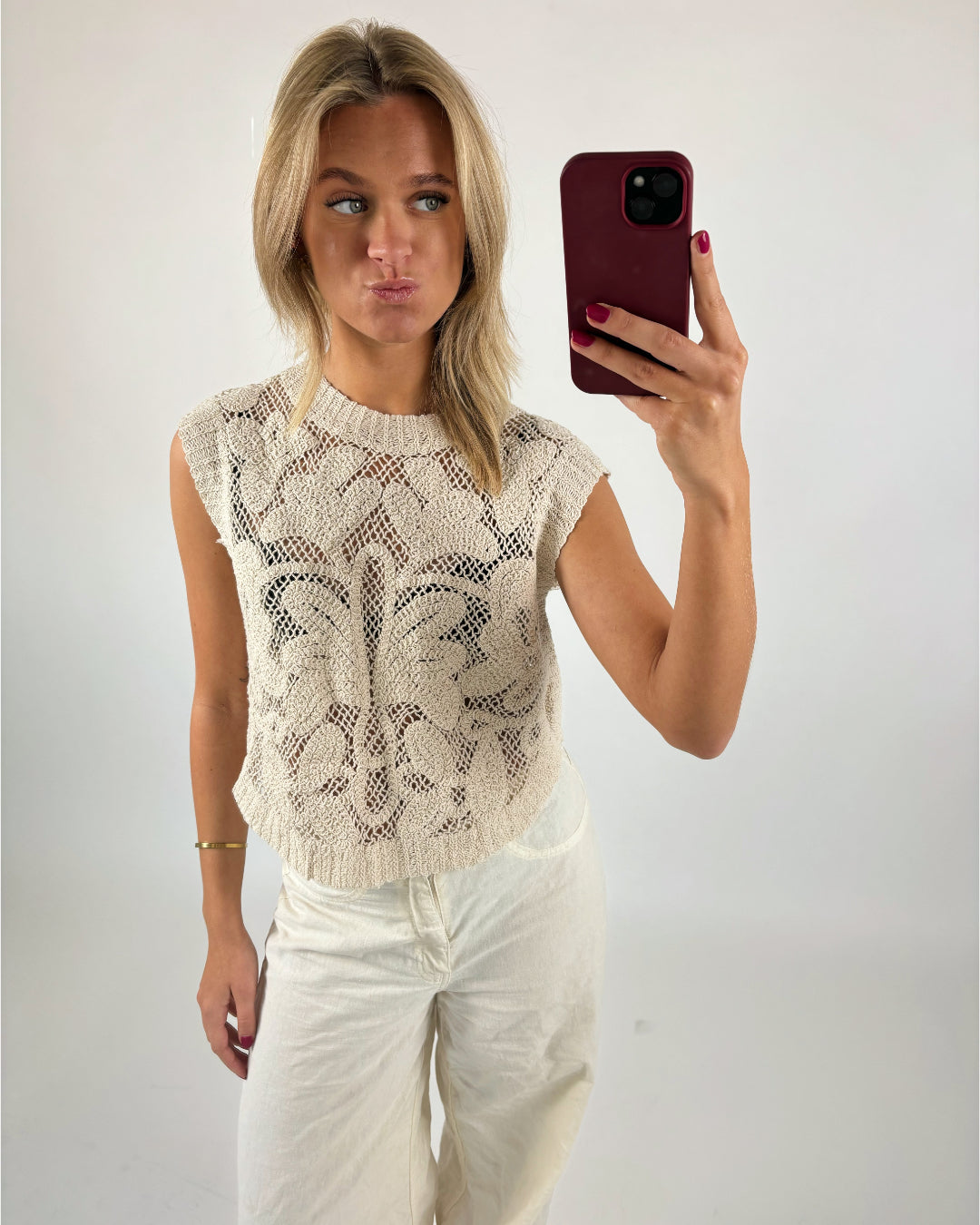 En person tager en selfie med en rød mobiltelefon iført en hvid, mønstret top og hvide bukser. Personen laver en sjov ansigt. Baggrunden er hvid og enkel.