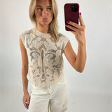 En person tager en selfie med en rød mobiltelefon iført en hvid, mønstret top og hvide bukser. Personen laver en sjov ansigt. Baggrunden er hvid og enkel.