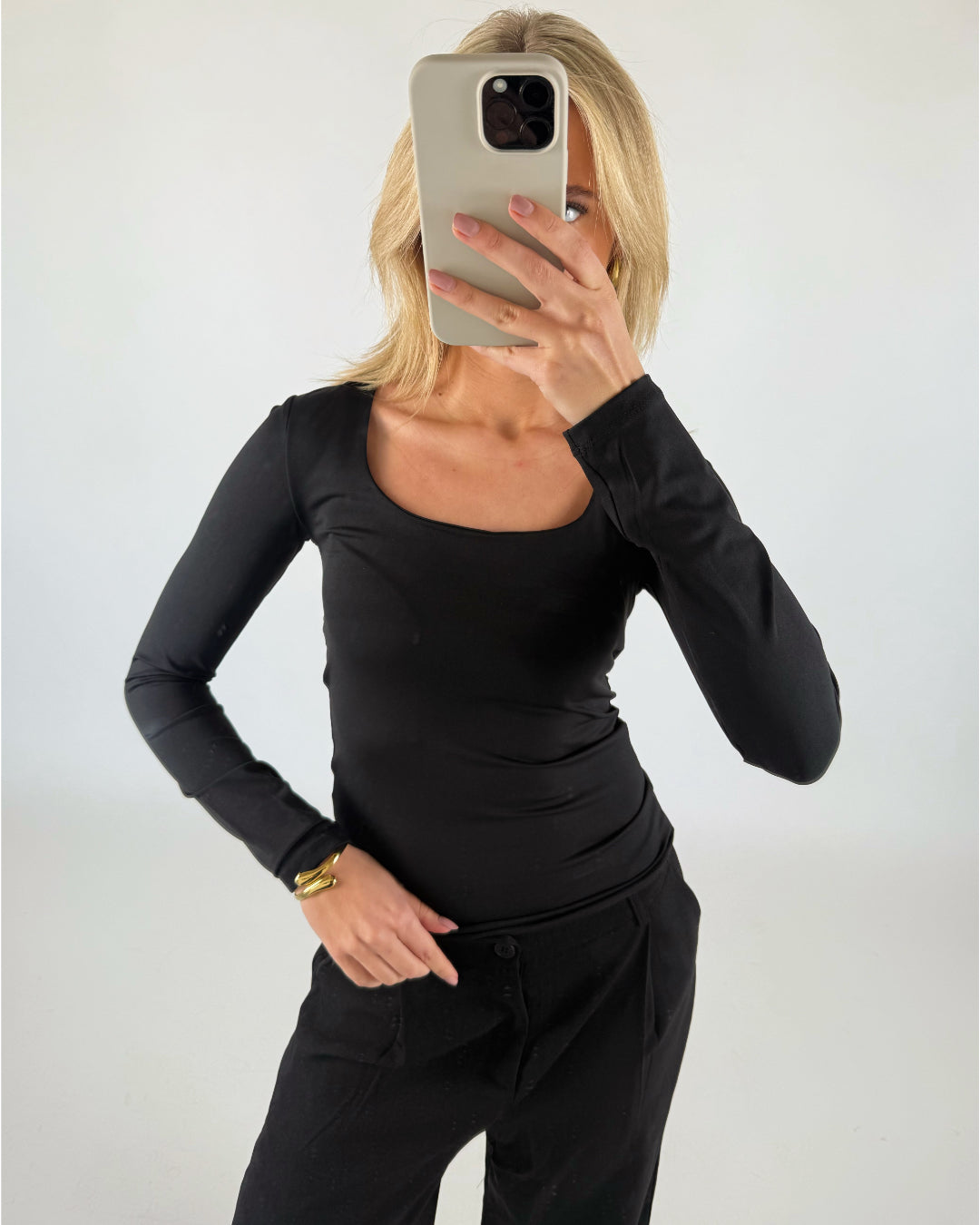 En person i sort skjorte og bukser tager et selfie foran en lys baggrund, hvor ansigtet er dækket af en mobiltelefon med et lysegråt cover.