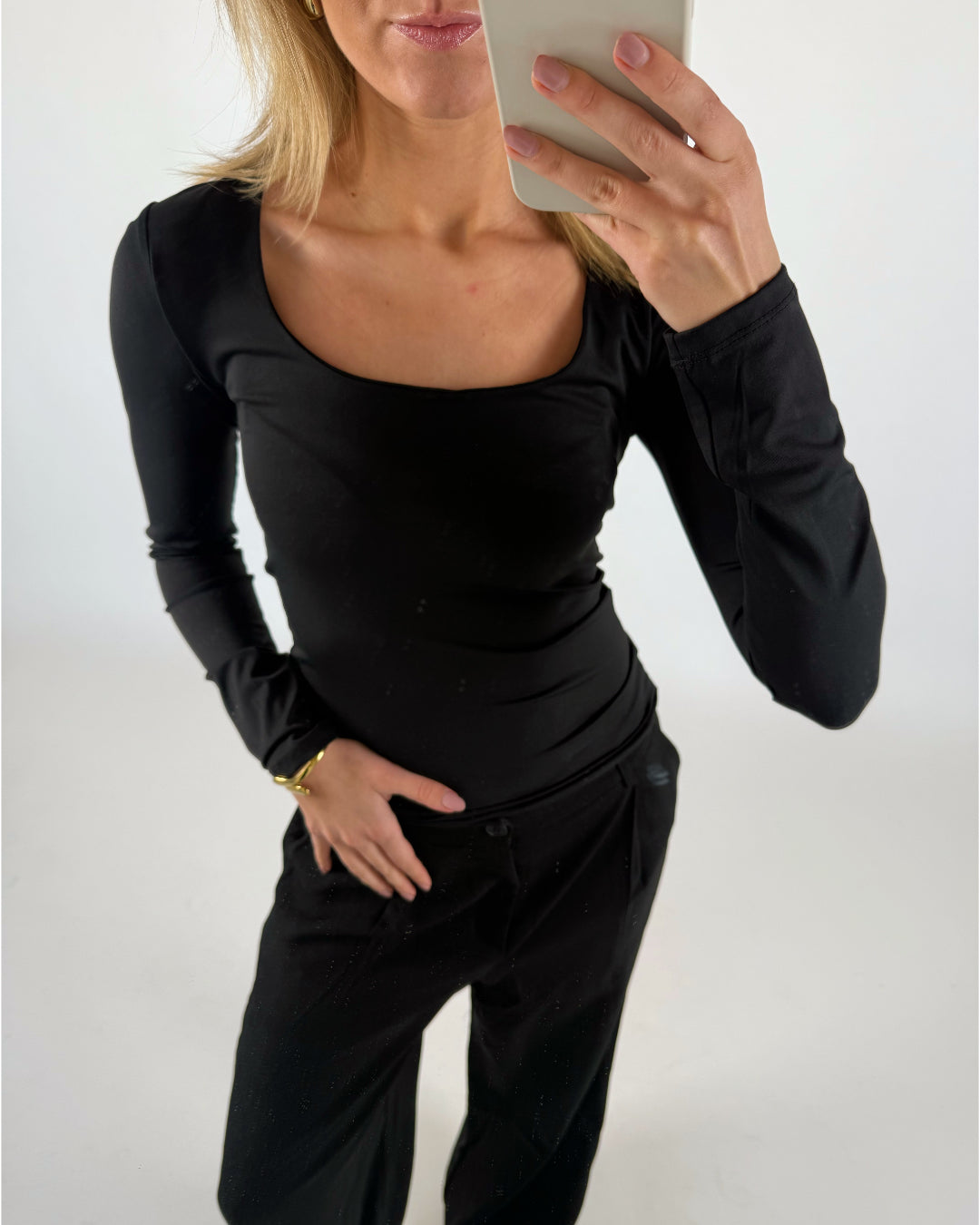 Person tager en selfie i spejl, iført sort trøje og bukser. Hånden holder en lysegrå telefon, mens en lys, minimalistisk baggrund opdager. Venstre hånd er på hoften.