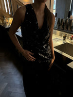 En person iført en sort, glitrende top står ved en bar med metaloverflader og cocktailshakere i baggrunden. Skiltet "Negroni" ses svagt på væggen.