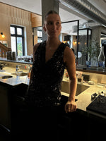 En person iført en glitrende sort top står ved en bar, omringet af glas og flasker, i en stilfuld café med dæmpet belysning og moderne indretning.