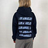 En person står med ryggen til iført en sort hættetrøje med teksten "LOS ANGELES" gentaget fem gange i hvid tekst. Baggrunden er lys og ensfarvet.