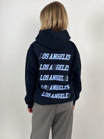 En person står med ryggen til iført en sort hættetrøje med teksten "LOS ANGELES" gentaget fem gange i hvid tekst. Baggrunden er lys og ensfarvet.