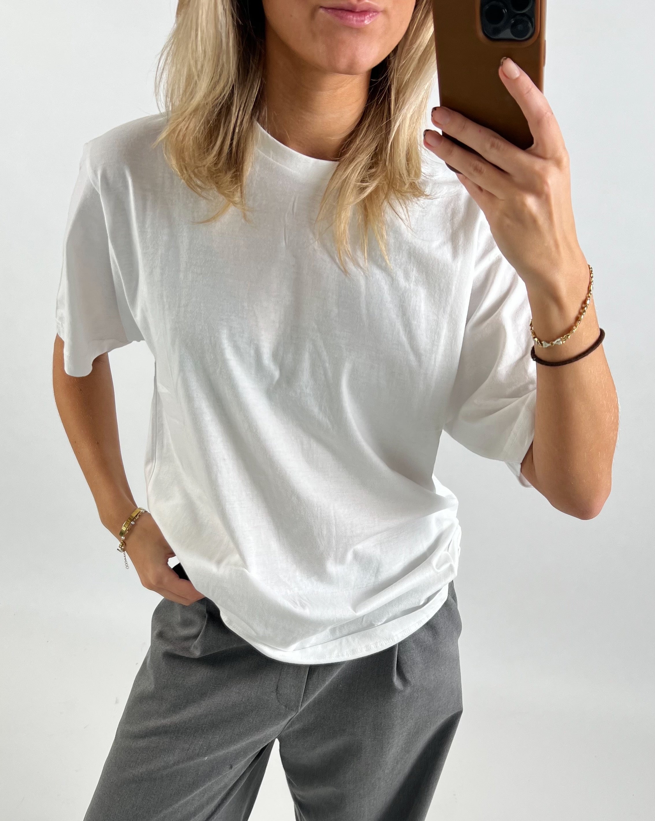 Person iført hvid t-shirt og grå bukser tager et selfie med brun mobiltelefon foran en neutral baggrund. Hånden i bukselommen, anden hånd holder telefonen.