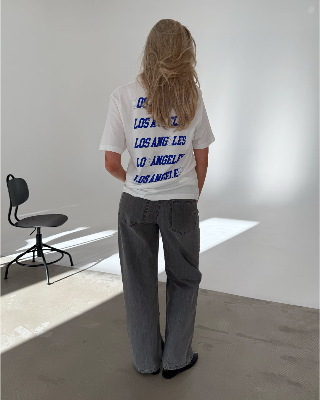 En person med langt hår iført en hvid T-shirt med teksten "LOS ANGELES" flere gange på ryggen, står i et lyst rum med en sort stol ved siden af.