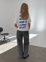 En person med langt hår iført en hvid T-shirt med teksten "LOS ANGELES" flere gange på ryggen, står i et lyst rum med en sort stol ved siden af.