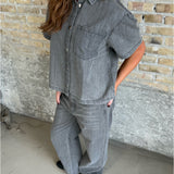 En person iført gråt denim outfit står afslappet mod en rå murstensvæg. Personens hænder er i lommerne, og de bærer sorte sko på et gråt betongulv.