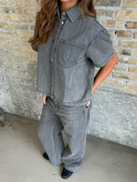 En person iført gråt denim outfit står afslappet mod en rå murstensvæg. Personens hænder er i lommerne, og de bærer sorte sko på et gråt betongulv.