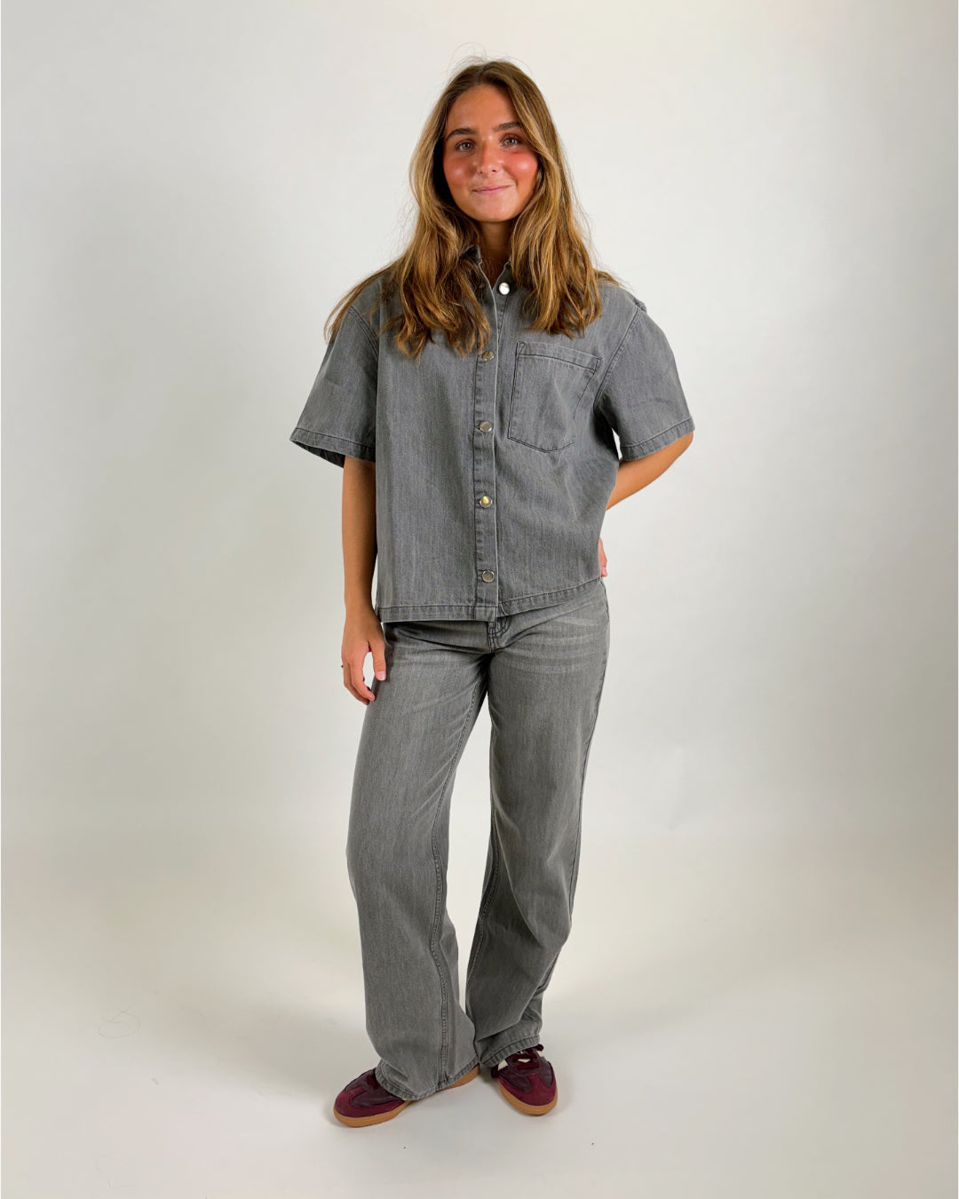 En person, iført en grå denimskjorte og matchende bukser, står afslappet med hænderne på hofterne. Baggrunden er enkel og lys.