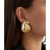 Ørering, skinnende og bølgende, hænger fra øret. Personen har mørkt hår og bærer en sort top, hvilket fremhæver smykkets gyldne glans i en elegant stil.