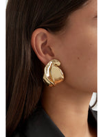 Ørering, skinnende og bølgende, hænger fra øret. Personen har mørkt hår og bærer en sort top, hvilket fremhæver smykkets gyldne glans i en elegant stil.