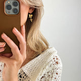 En person holder en mobiltelefon og ser i spejlet, iført en hvid strikket trøje og gyldne øreringe. Baggrunden er minimalistisk og lys.