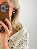 En person holder en mobiltelefon og ser i spejlet, iført en hvid strikket trøje og gyldne øreringe. Baggrunden er minimalistisk og lys.