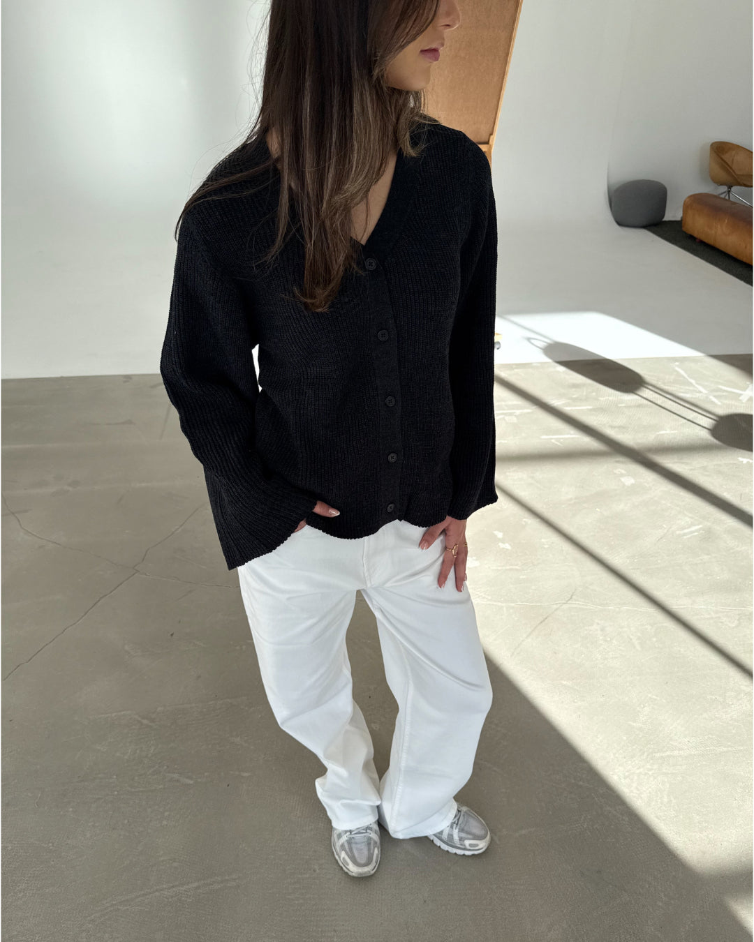 En person står i en sort cardigan og hvide bukser, ser mod siden, i et lyst rum med en brun sofa.