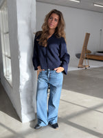 Kvinden står afslappet op ad en hvid mur, iført en mørk trøje og lyse jeans. Rummet har store vinduer og moderne indretning med minimalisme som stil.
