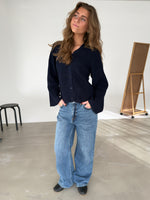 En person står afslappet med hænderne i lommerne iført en mørk striktrøje og blå jeans. Baggrunden inkluderer en skammel og et staffeli i et lysegråt rum.