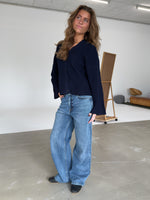 En person i en mørk bluse og brede jeans står afslappet i et rummeligt, minimalistisk rum med en lysebrun sofa og en staffeli i baggrunden.