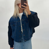 En person iført en denimjakke og lys denimnederdel tager et spejlbillede med en smartphone i et neutralt rum.