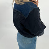 En person står iført en sort og blå jakke og lyst hår, tager et spejlbillede med en smartphone. Personen bærer også en lyseblå denim nederdel. Baggrunden er hvid.