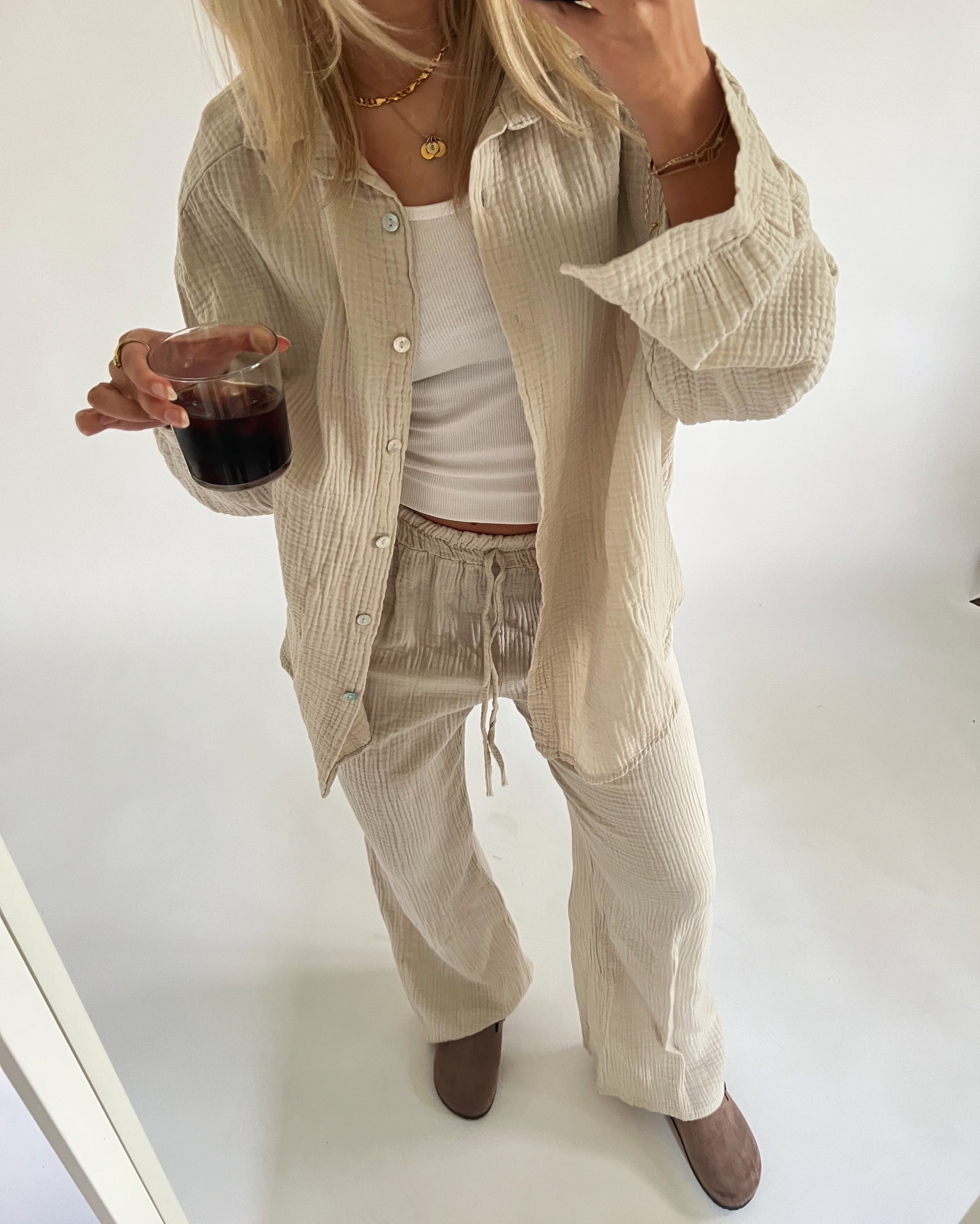 En person iført beige sæt drikker fra et glas. Personen står i et lyst rum med hvid baggrund og brune sko.