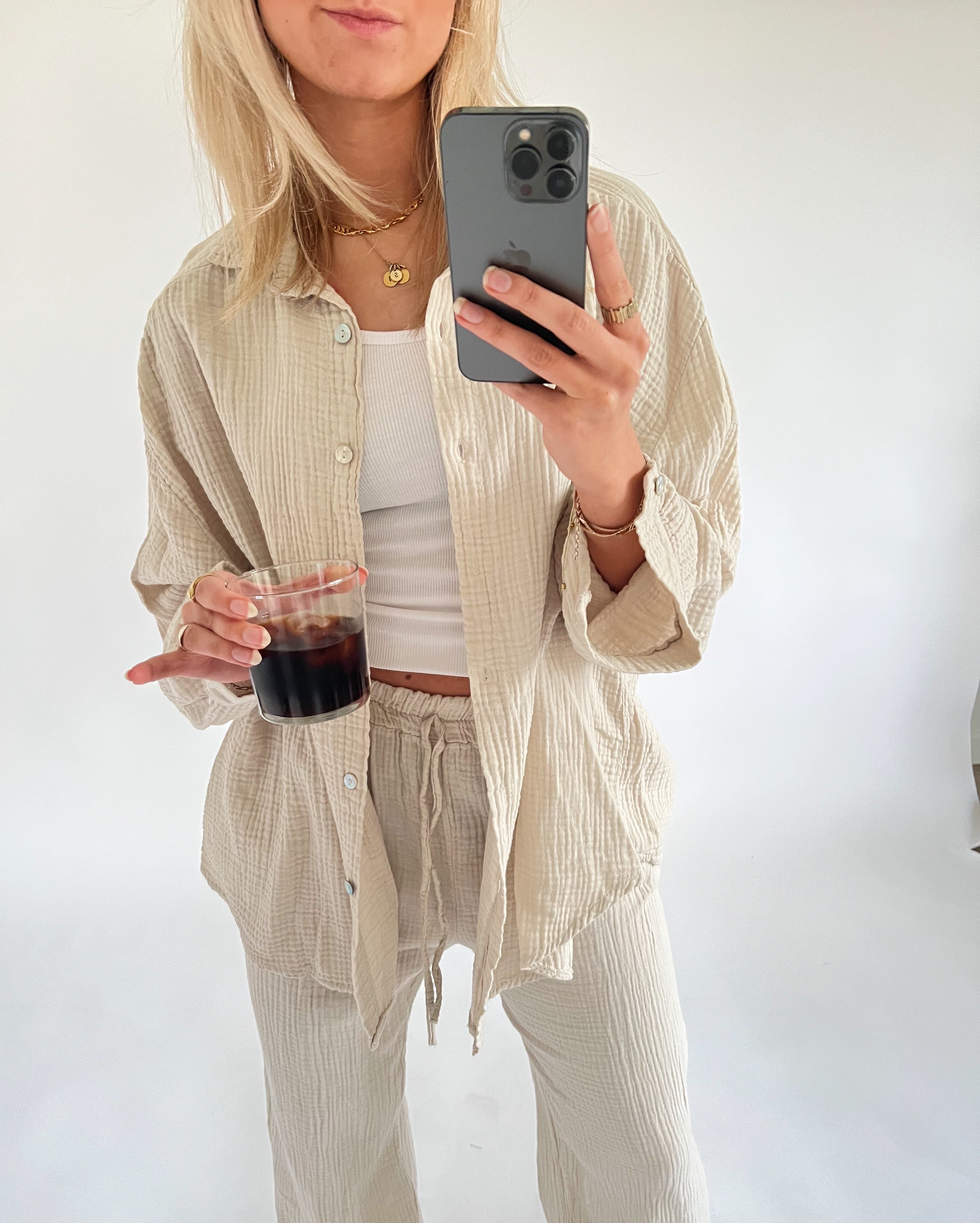 En person iført lys, løs bomuldsskjorte og bukser holder en smartphone og et glas mørk væske. Personen står i et lyst rum uden synlig tekst.