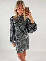 En person i en skinnende, sølvfarvet kjole tager en selfie med en smartphone i et neutralt rum og laver en kysmund.