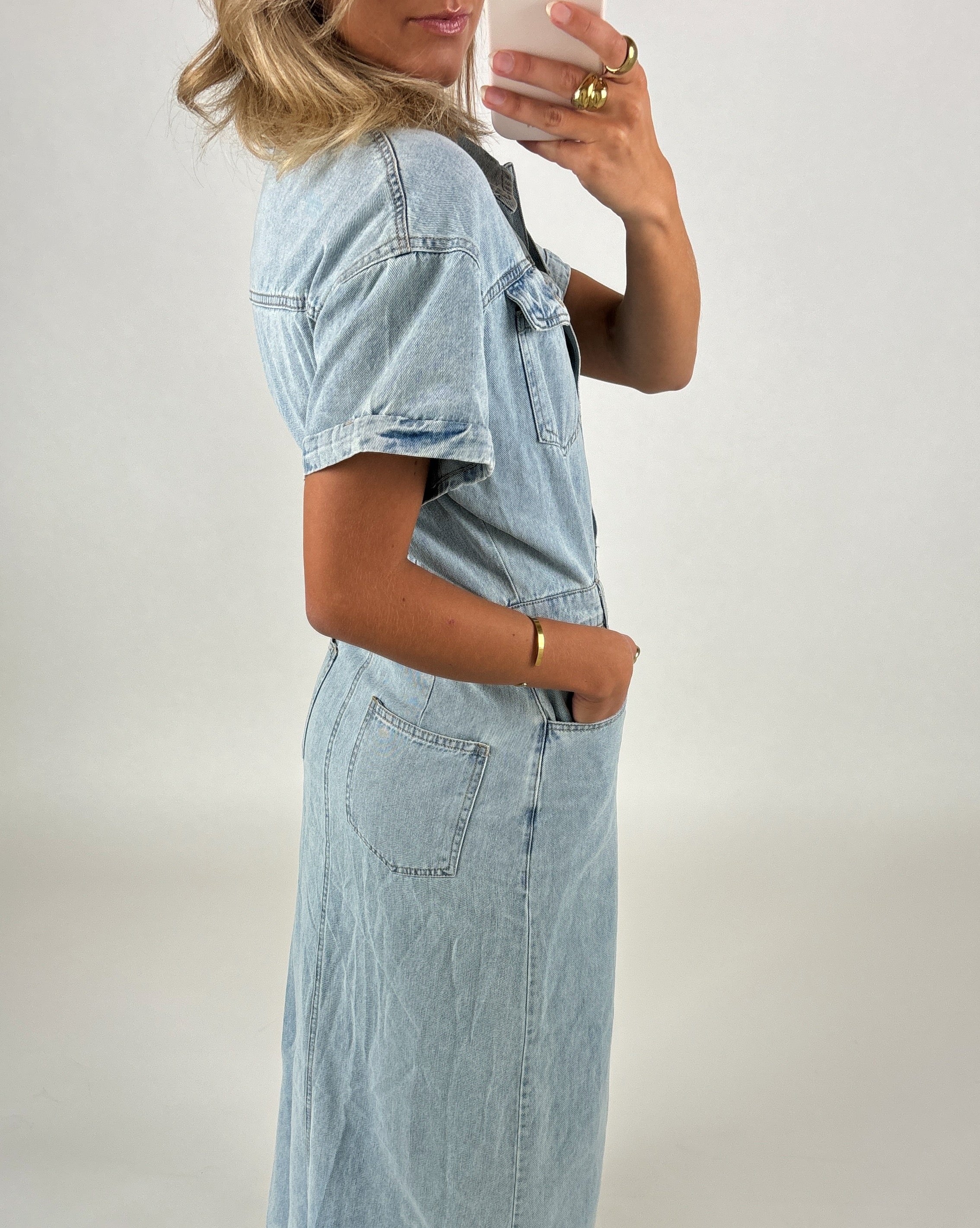 En person i en lyseblå denimkjole står sidelæns med en hånd i lommen og den anden løftet, mens de holder en telefon i et minimalistisk, neutralt rum.