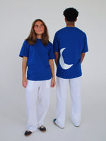 To personer iført blå T-shirts, en vender fremad og en med en halvmåne på ryggen, i et lyst rum iført hvide bukser.