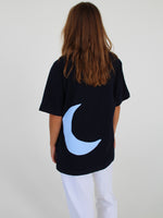 En person iført en sort T-shirt med en blå måne på ryggen står med ryggen til og har langt, brunt hår. De har hvide bukser på i et neutralt rum.