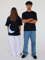 To personer står i et lyst rum. Den ene bærer en sort t-shirt med en blå halvmåne og hvide bukser, vendt bagud. Den anden har en sort t-shirt, krydsede arme og jeans.