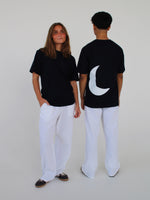 To personer iført sorte T-shirts og hvide bukser står i et hvidt rum; den ene er vendt mod kameraet, mens den anden viser en hvid halvmåne på ryggen.