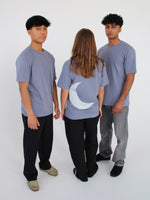Tre personer står sammen i en cirkel, iført grå t-shirts. En person vender ryggen til, med en måneform på trøjen. De er i et minimalistisk, hvidt rum.