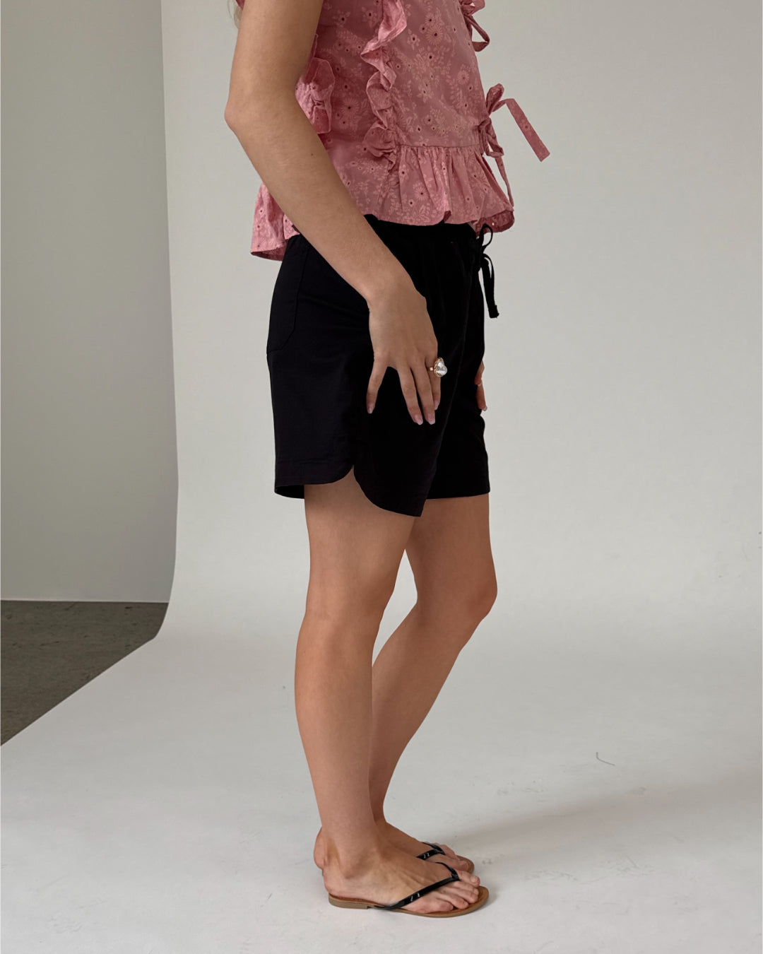 En person iført lyserød bluse og sorte shorts står på et hvidt og neutralt baggrundsgulv. Personen har sandaler og har en hånd hvilende langs kroppen.