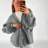 En person iført en grå, strikket cardigan tager en selfie med en telefon i en brun etui. Personen bærer en hvid t-shirt og hvide bukser i et lys rum.
