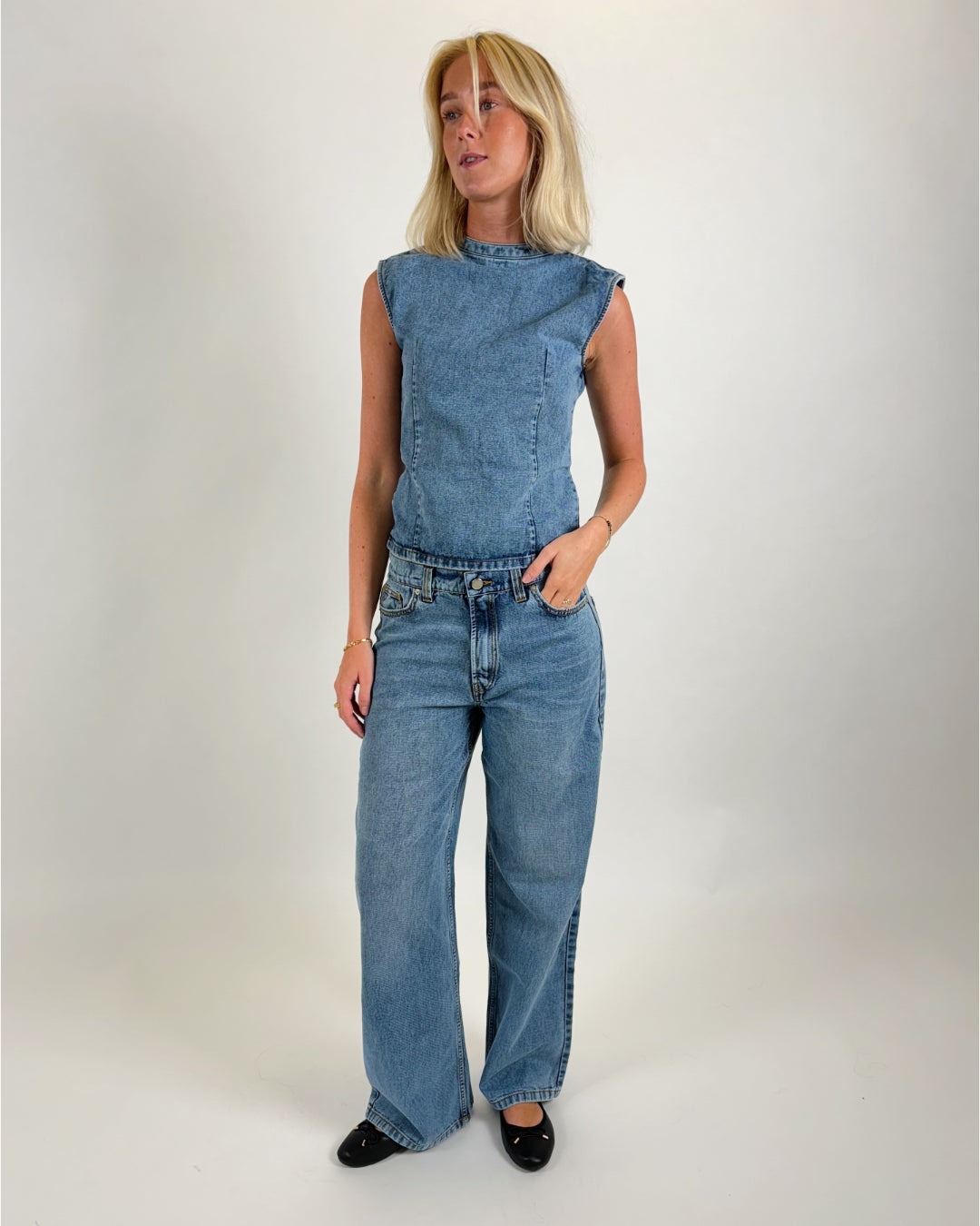 Kvinde iført ærmeløs denimtop og jeans, står med hånden i lommen i et minimalistisk, lyst studie, iført sorte sko.