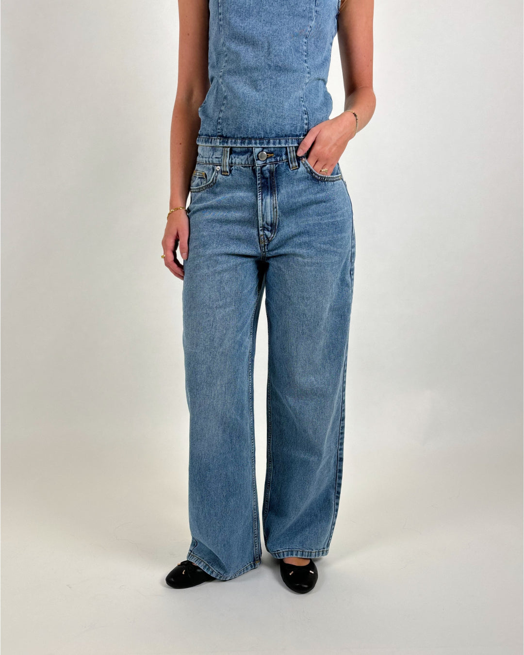 En person bærer en denim jumpsuit og sorte sko, står med en hånd i lommen i et neutralt, lyst miljø.