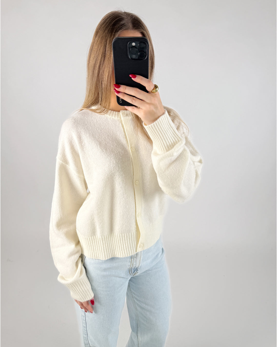 En person iført en cremefarvet cardigan tager et spejlbillede med en mobiltelefon. Personen har rødlakerede negle og en stor ring. Baggrunden er hvid og minimalistisk.