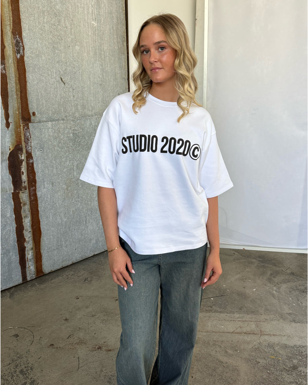 En person med blondt, krøllet hår står, iført en hvid T-shirt med teksten "STUDIO 2020©" og jeans. Baggrunden er rustikt med metaldetaljer og en minimalistisk væg.