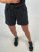 En person iført sorte shorts med mønster står med en hånd ved taljen. De bærer også sorte og gule sneakers på en hvid baggrund.