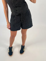 Person iført sort mønstret shorts og top, står på et hvidt gulv. De bærer sorte sneakers med gule striber og guldsmykker på håndleddet.